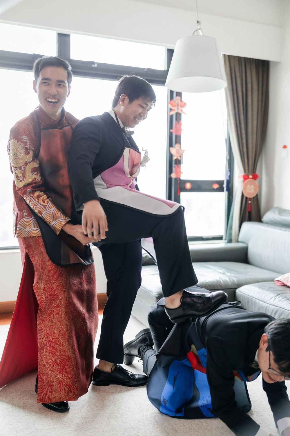 Groomsmen playfully helping groom dress in traditional Chinese wedding attire indoors.