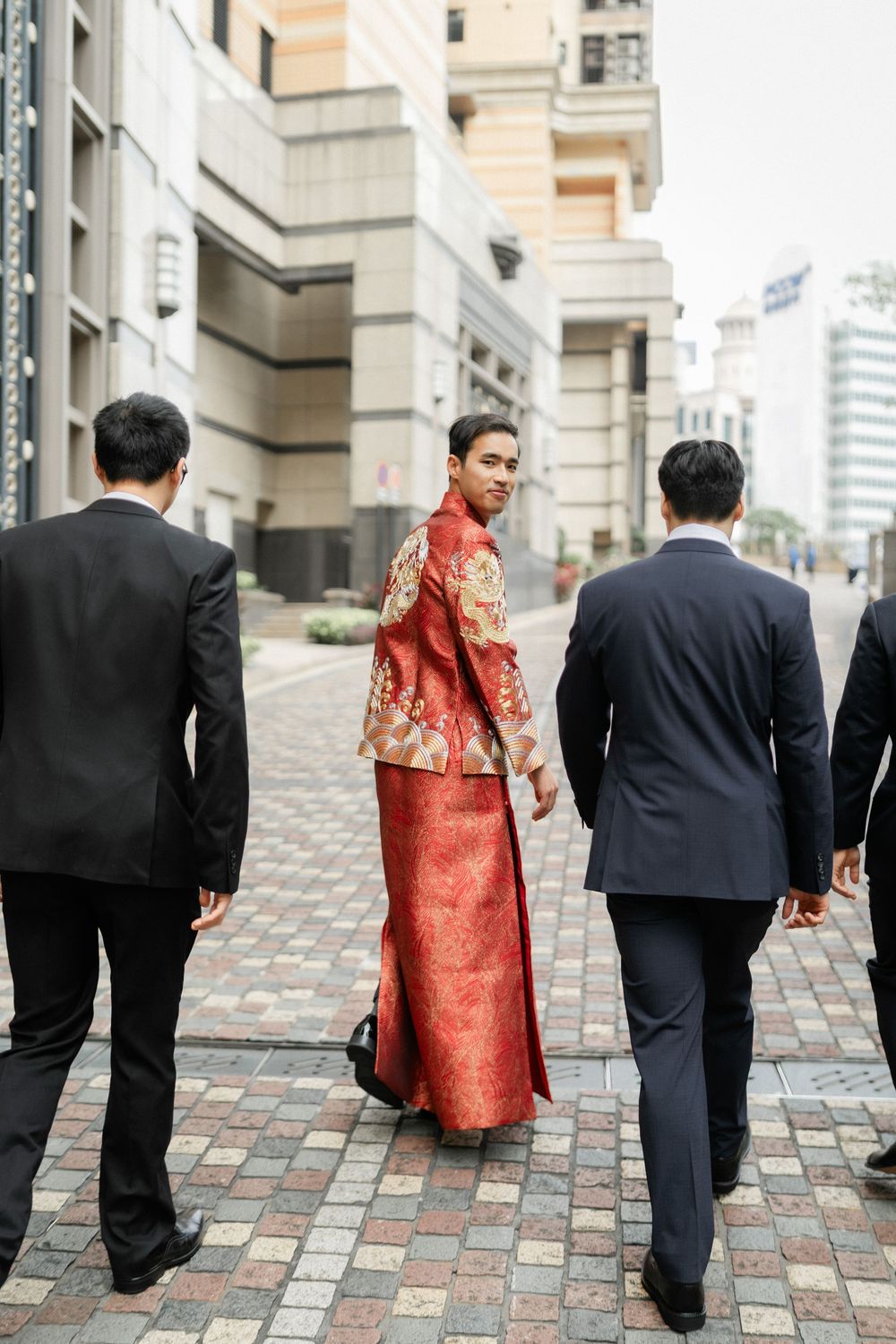 Groom in red Chinese traditional outfit walks with groomsmen in black suits along city street.