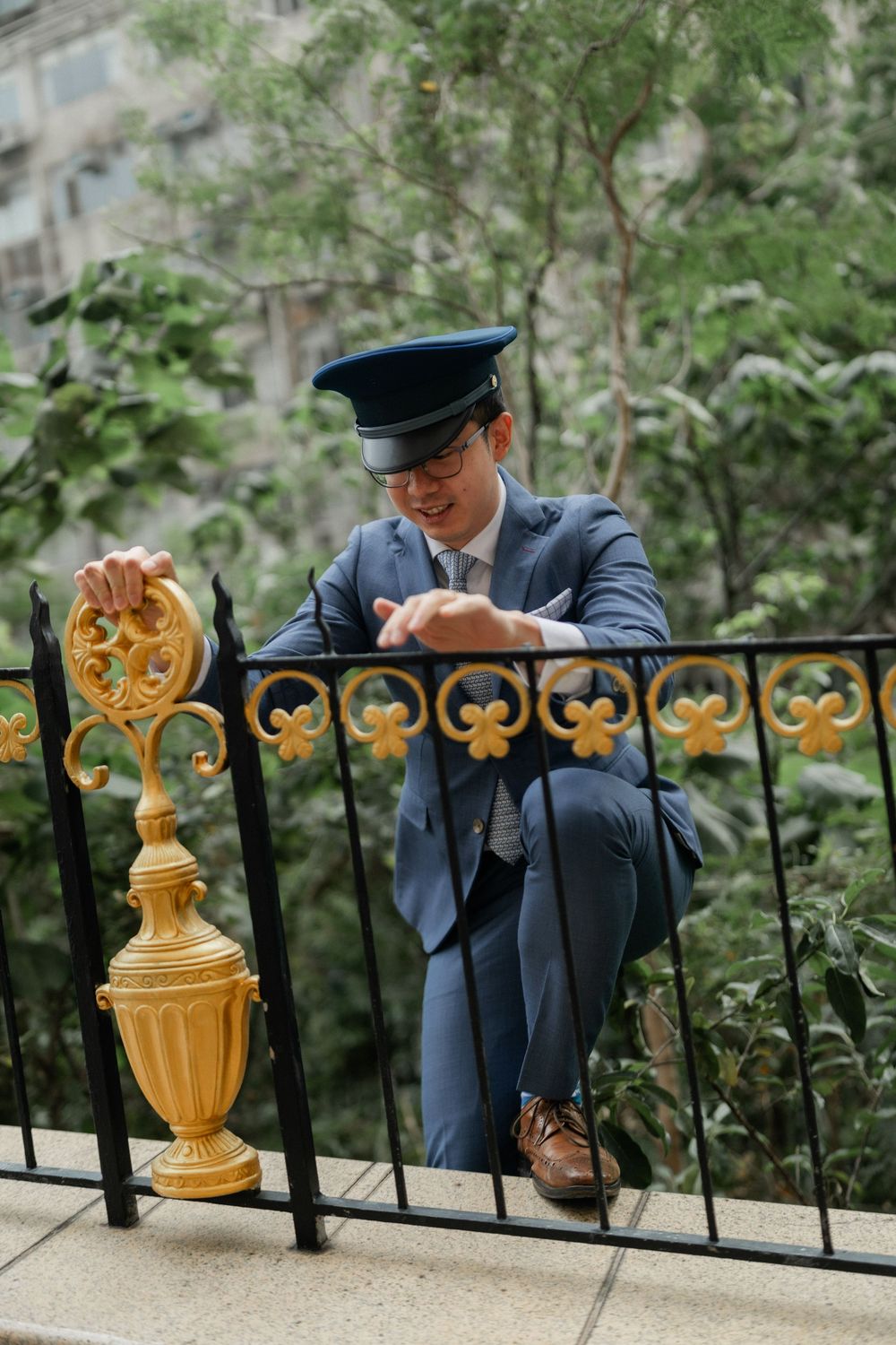 Man in blue suit and cap crouches by ornate black and gold iron fence outdoors.