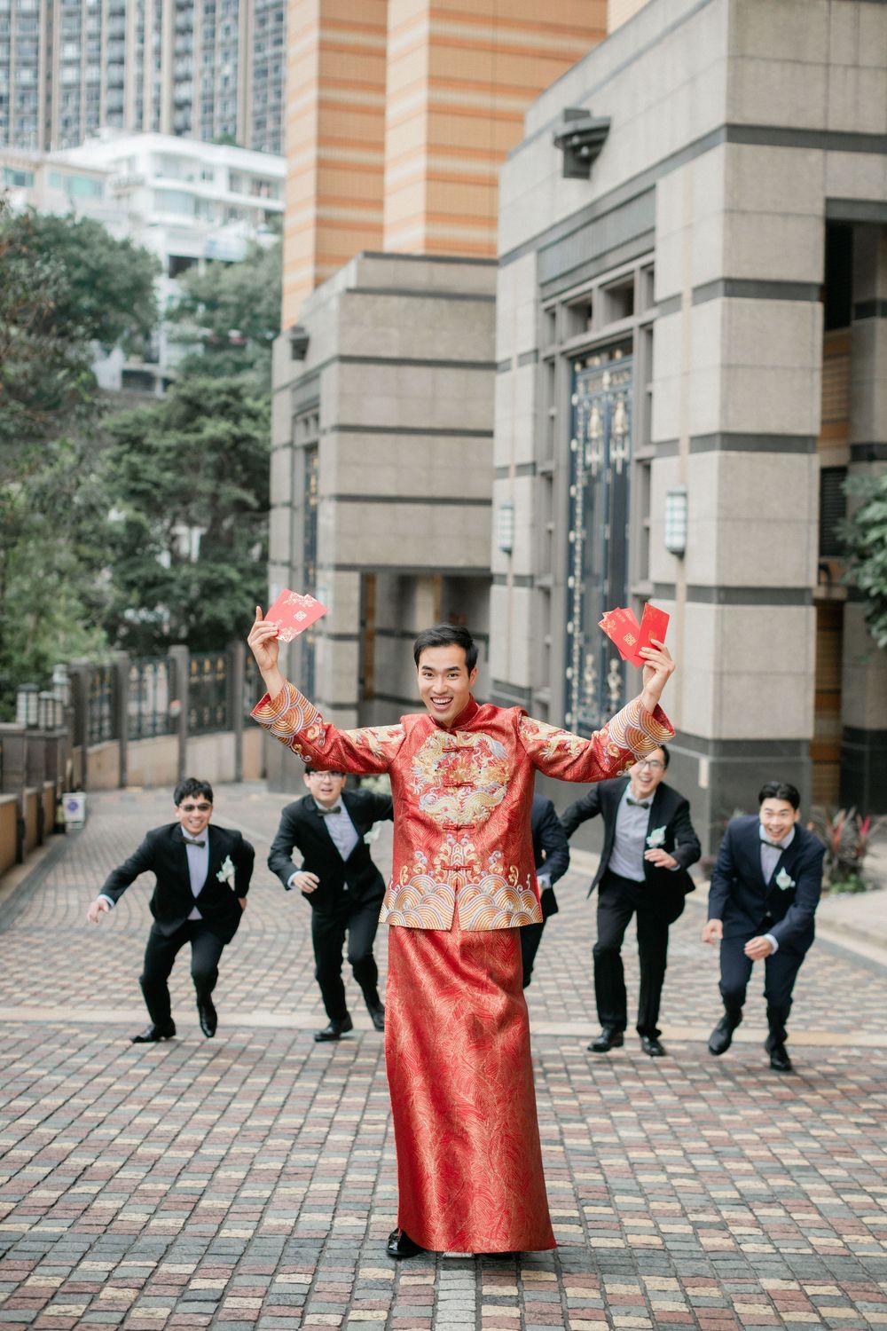 Groom in red Chinese wedding attire celebrates with red envelopes as groomsmen run toward him.