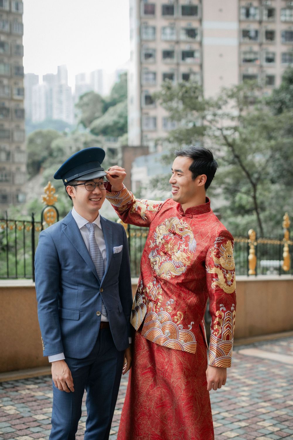 Groom in red Chinese traditional attire playfully adjusts cap on friend in blue suit outdoors.