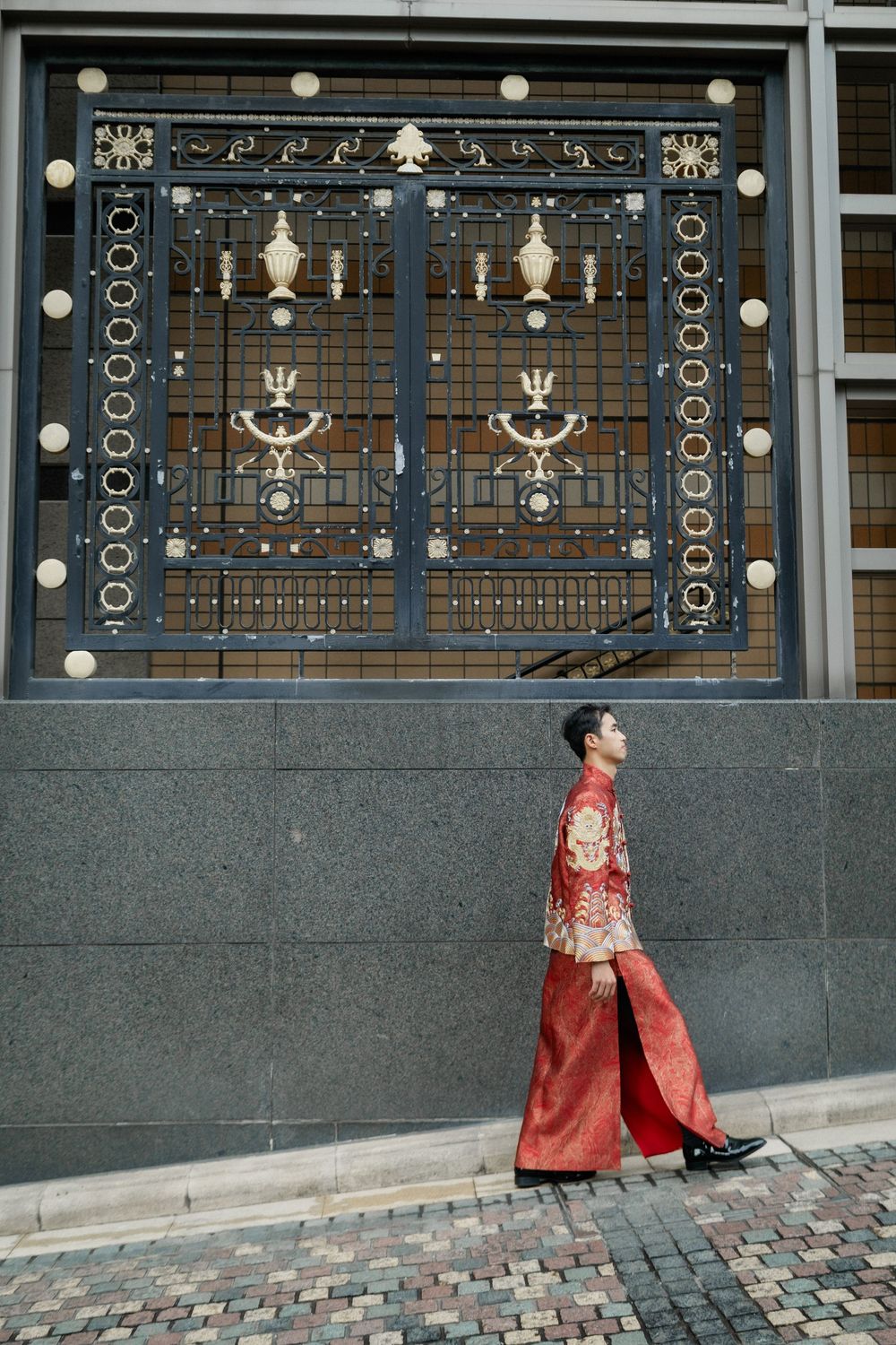 Groom in traditional red Chinese wedding robe walks past ornate decorative iron gate on street.
