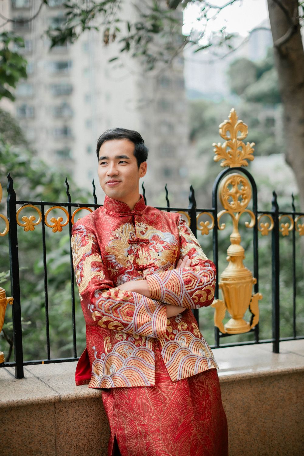 Groom in red embroidered Chinese wedding attire stands arms crossed by ornate golden fence.
