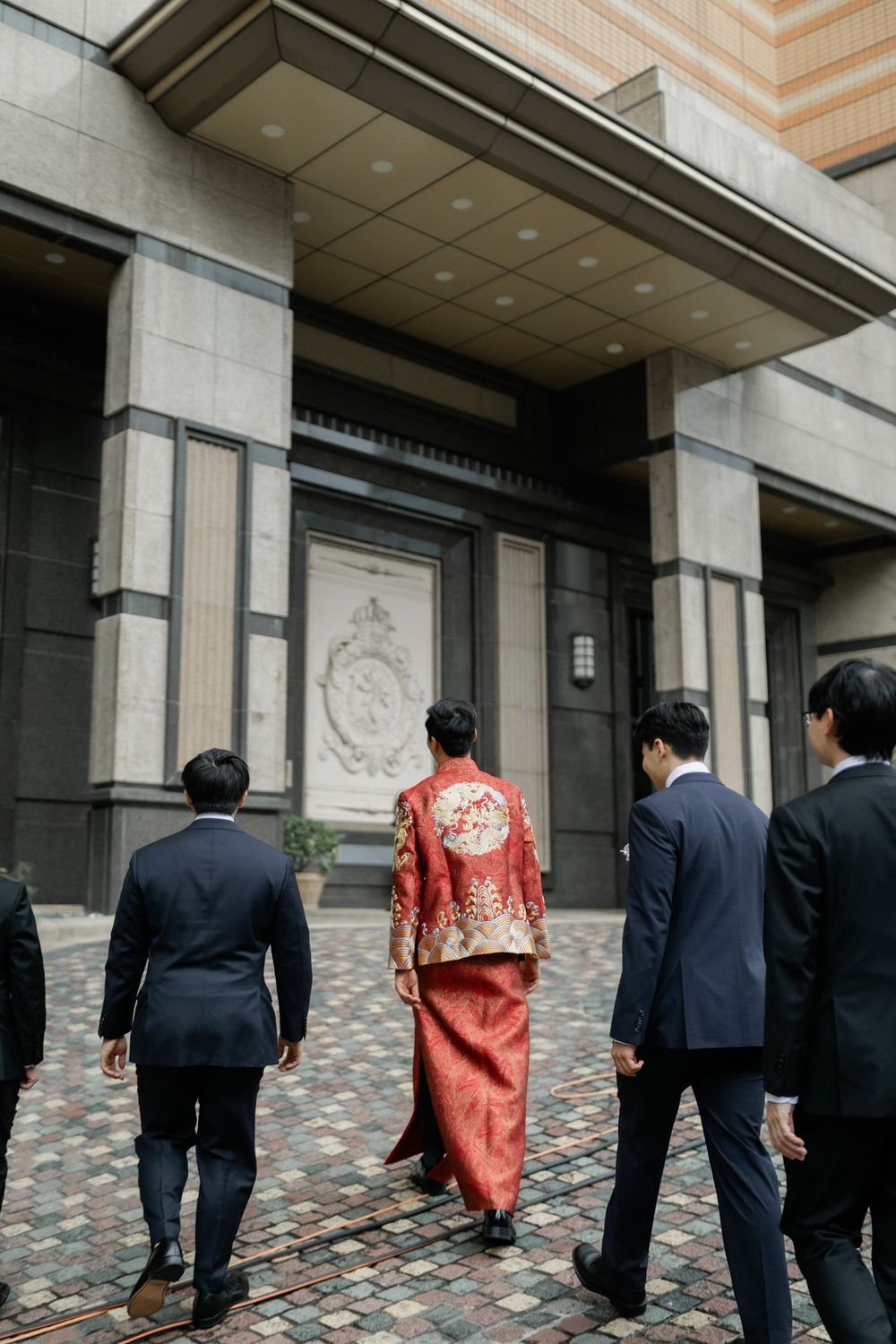 Groom in red Chinese traditional wedding robe enters building with groomsmen in black suits.