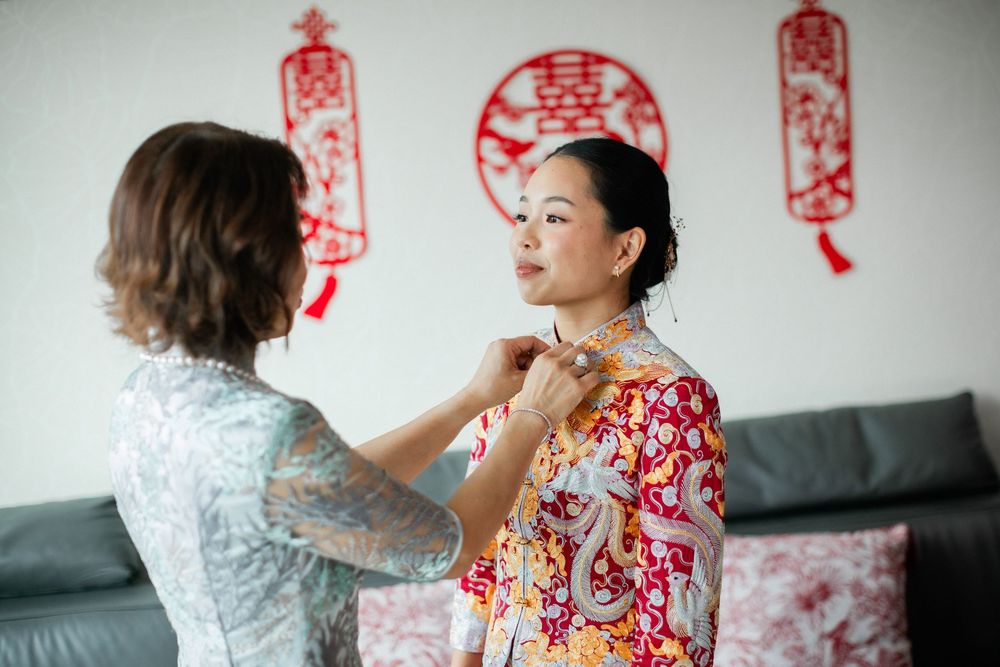 Woman helps bride in colorful floral qipao adjust collar with double happiness decorations behind.