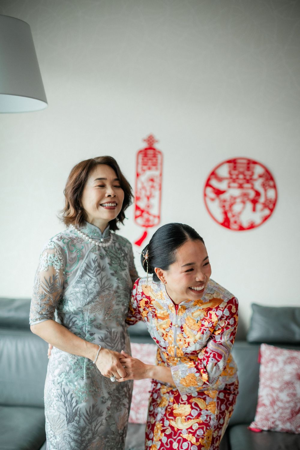 Two women in traditional Chinese qipao dresses laugh together with red double happiness wall decor.