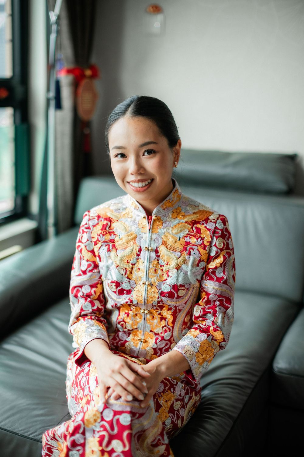 Smiling bride in ornate red and gold Chinese wedding qipao sitting on a sofa.