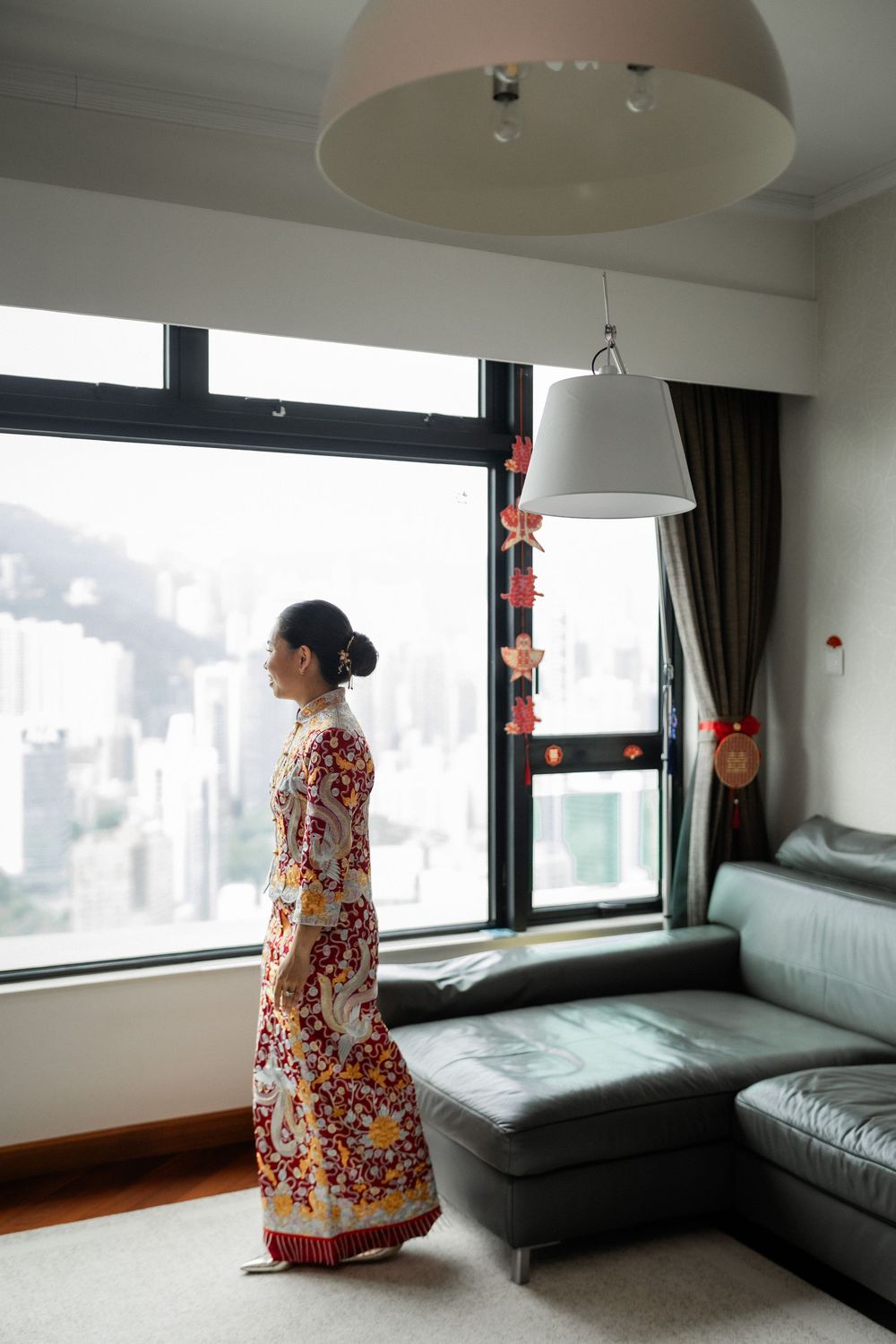 Bride in traditional Chinese qipao gazing out a window at a city skyline.