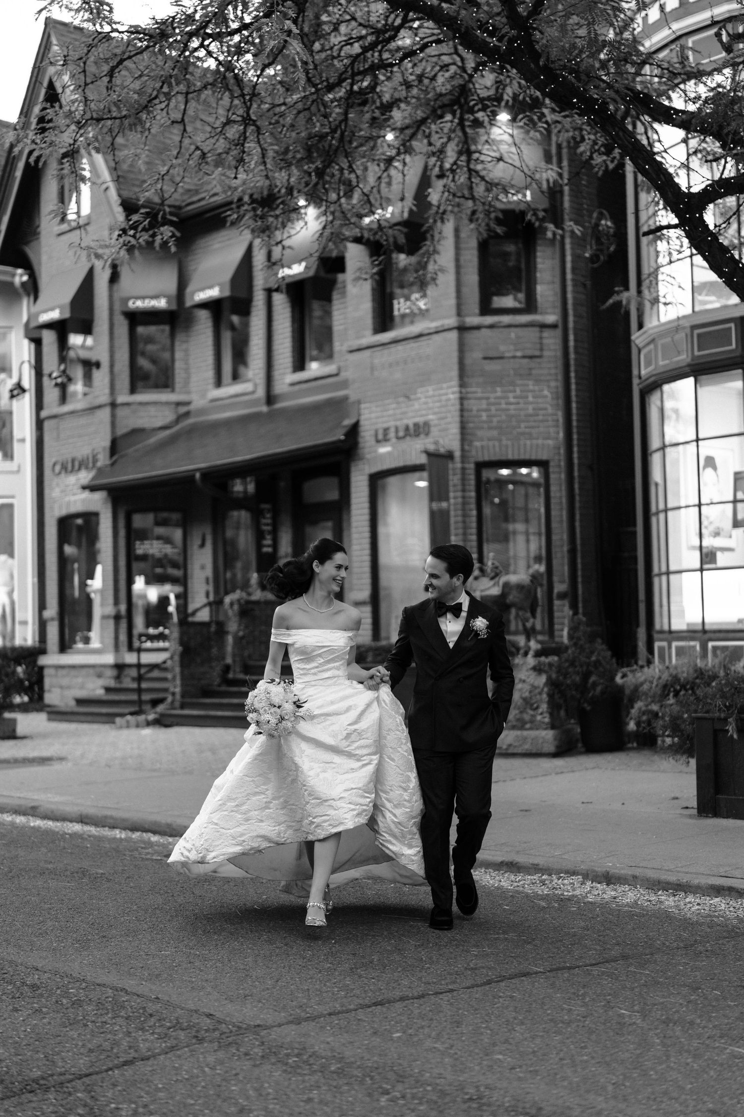 A romantic black and white wedding photo on a busy city street with tall buildings and a parked bicycle in the background.