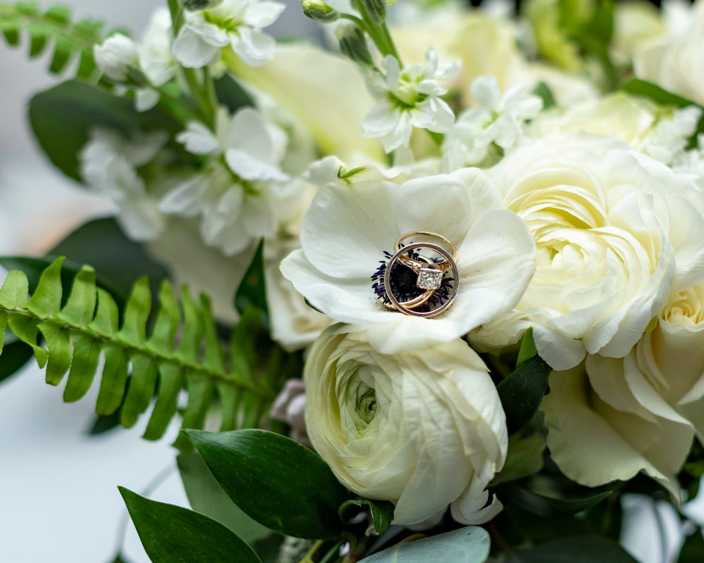 A delicate white floral arrangement with ranunculus, roses, and green ferns creates an elegant botanical display wedding rings in Concord.