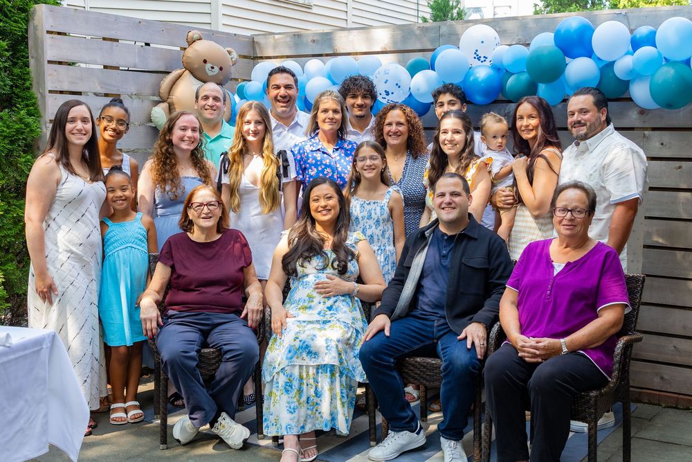 A large family group gather with mom and dad to be at their baby shower with blue and white balloons decorating the background in Boston.