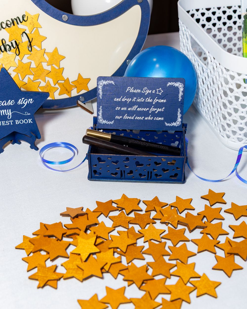 Wooden stars for well wished are scattered on white surface with blue guest book and decorative box in navy and white theme.