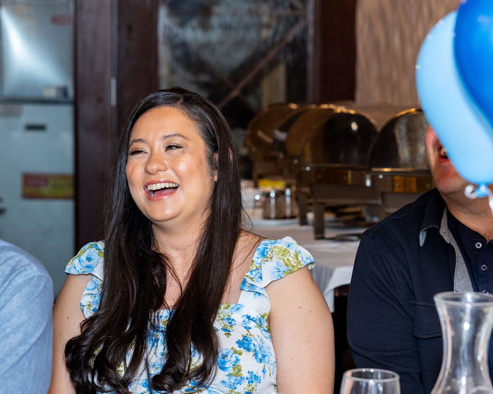 Mom to be in a floral dress laughs joyfully at her baby shower with blue balloons in the background.