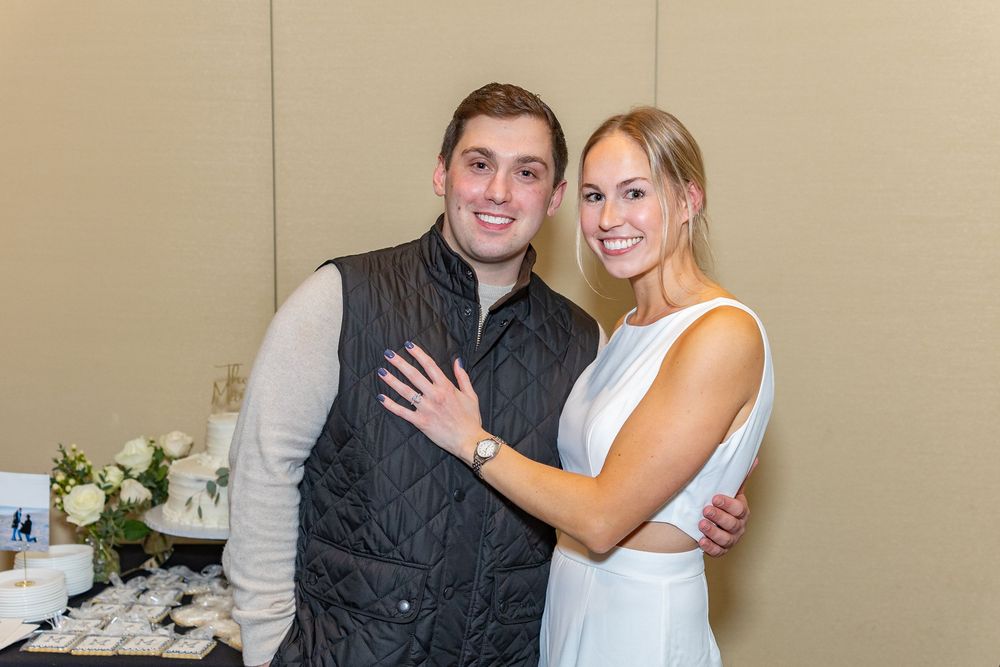 A couple embraces and smiles while standing against a beige wall at a formal event at their engagment party.