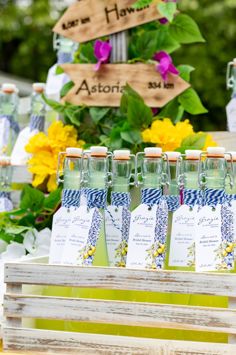 Glass bottles with blue and white checkered labels arranged in a row at an outdoor garden party setting at an Italian themed bridal shower.