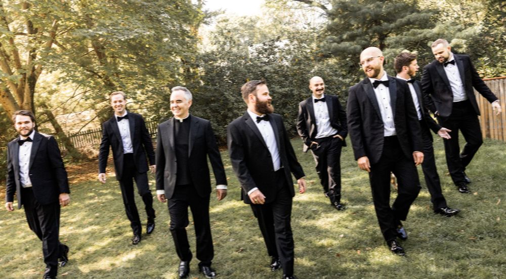 Groomsmen in black tuxedos walk together outdoors along a tree-lined path during autumn.