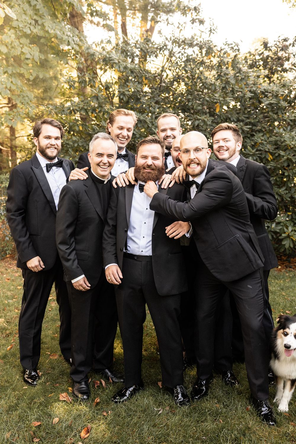 Groomsmen in black tuxedos pose playfully outdoors with a dog.