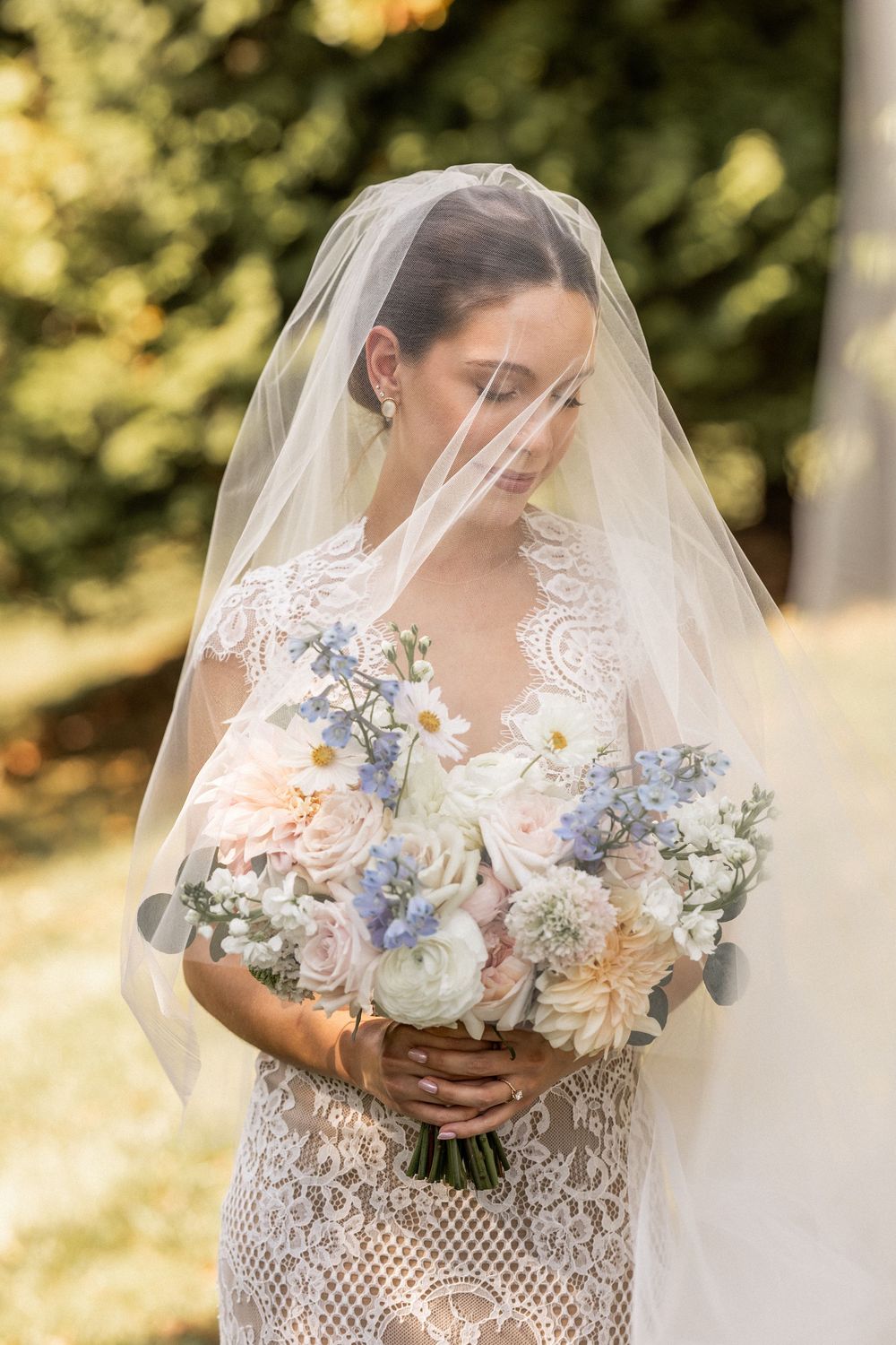 Bride in lace dress holding a pastel bouquet with blue and pink flowers against a sunlit outdoor backdrop.