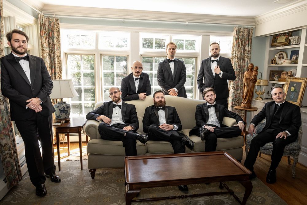 Groomsmen in black tuxedos sitting casually in a traditional living room setting.