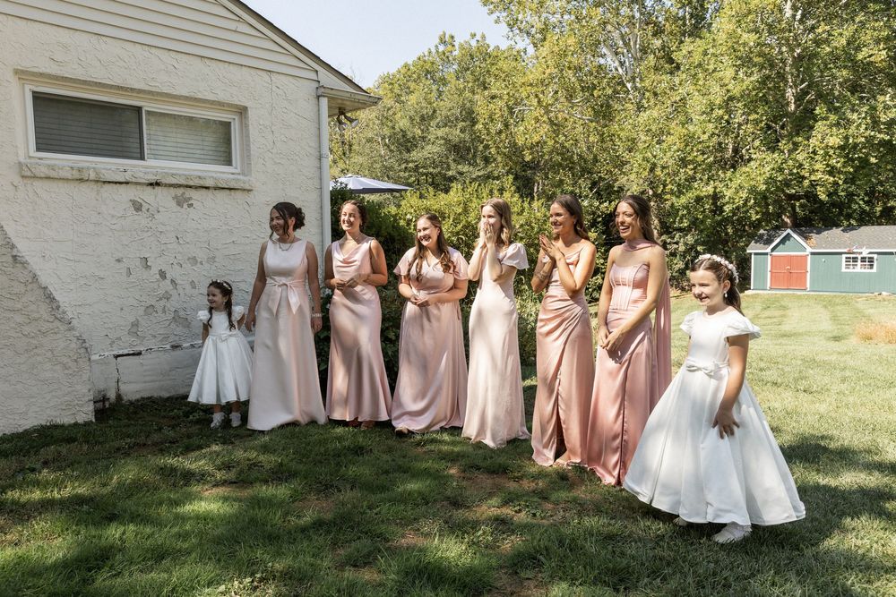 Wedding party in pink bridesmaid dresses and white gowns pose together outdoors in front of rustic building.
