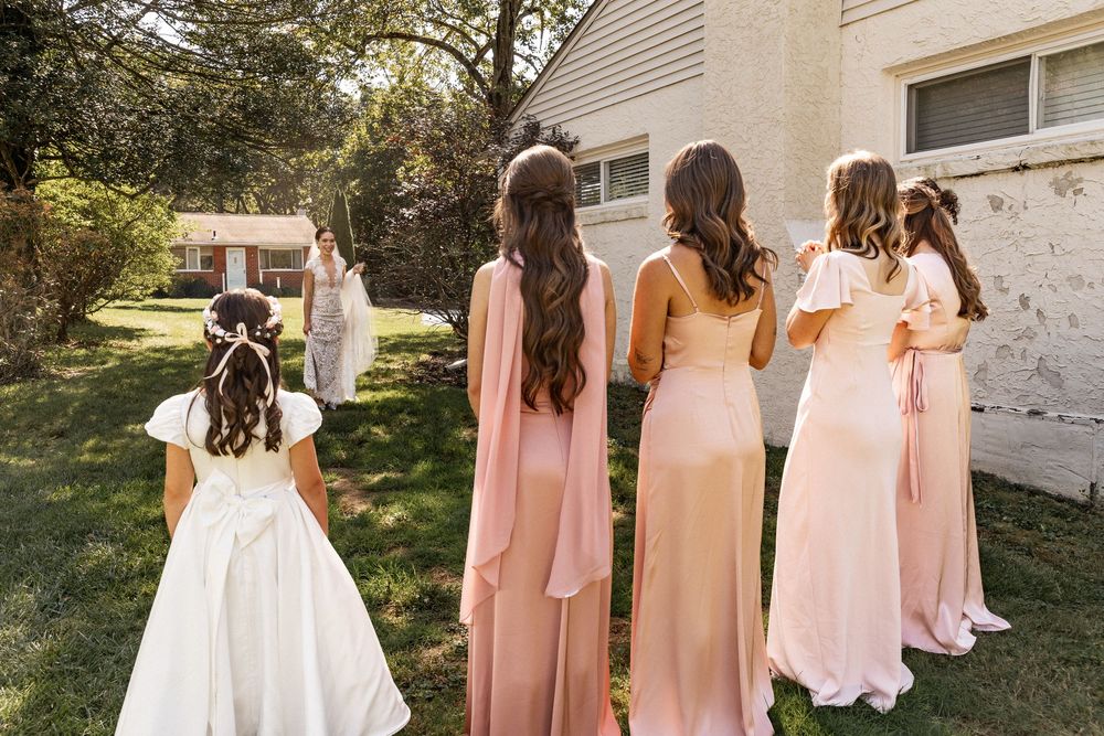 Wedding party members in blush pink dresses walk together on a sunny day outdoors.