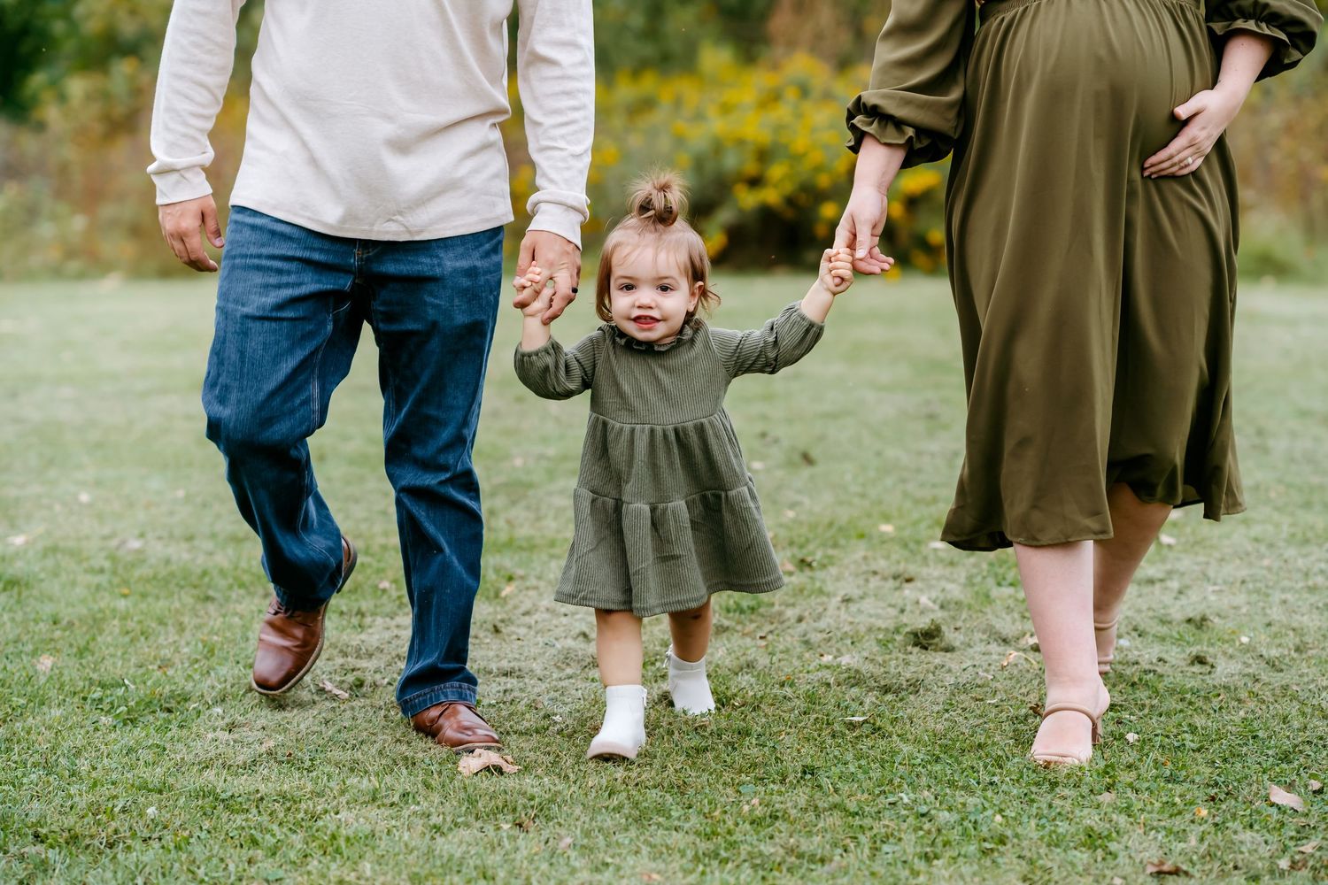 A family walking together on green grass, showing only their legs and feet in casual attire.