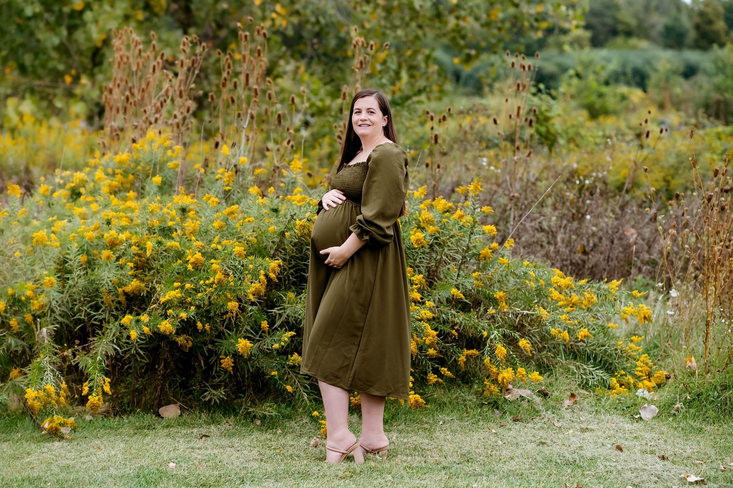 A woman in an olive green dress stands among vibrant yellow wildflowers in a lush meadow.