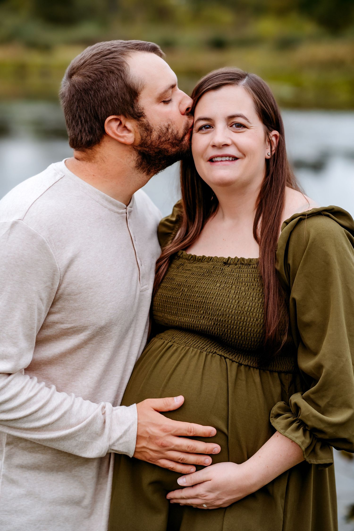 Intimate outdoor maternity photoshoot featuring a couple in coordinating olive and beige sweaters by a lake.