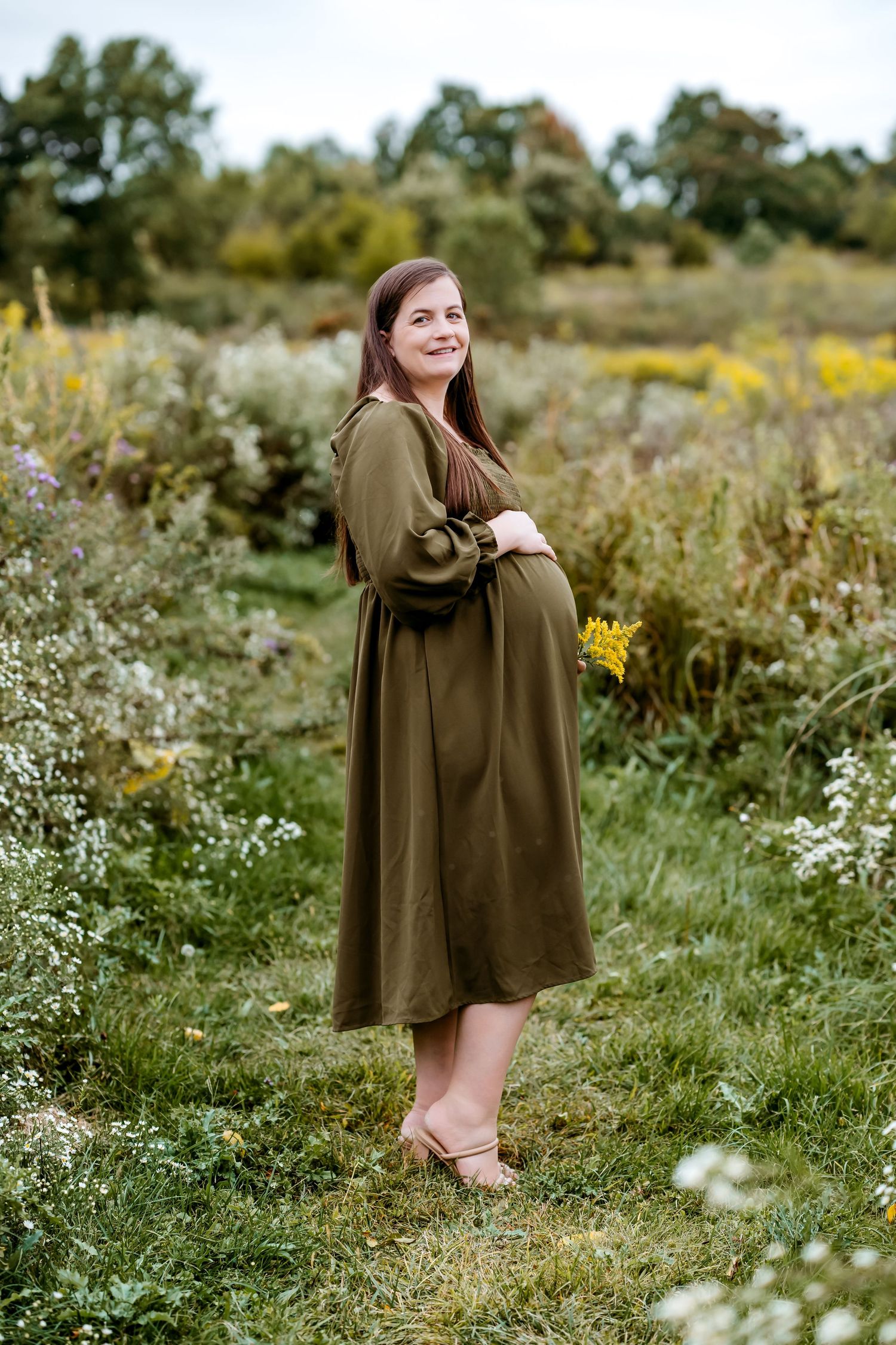 A person in an olive green cape poses in a wildflower meadow during a sunny outdoor photo session.