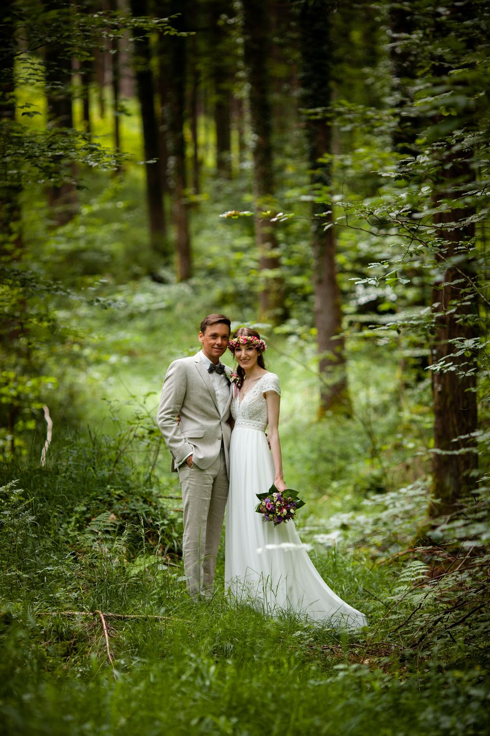 A romantic wedding couple walks through a lush green forest setting in elegant attire.