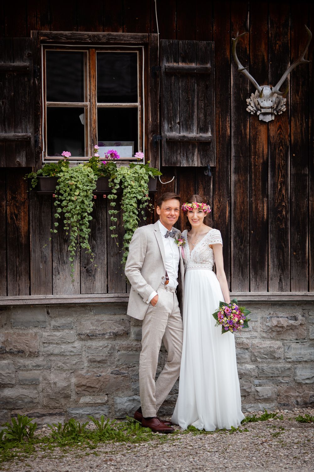 A couple in wedding attire poses against a rustic wooden barn wall with stone foundation and window boxes.