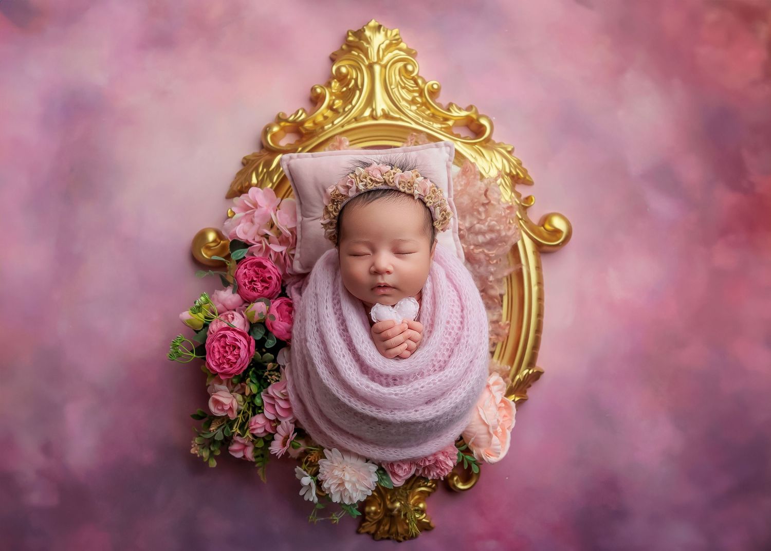 Sleeping newborn wrapped in pink swaddle inside an ornate gold frame with flowers.