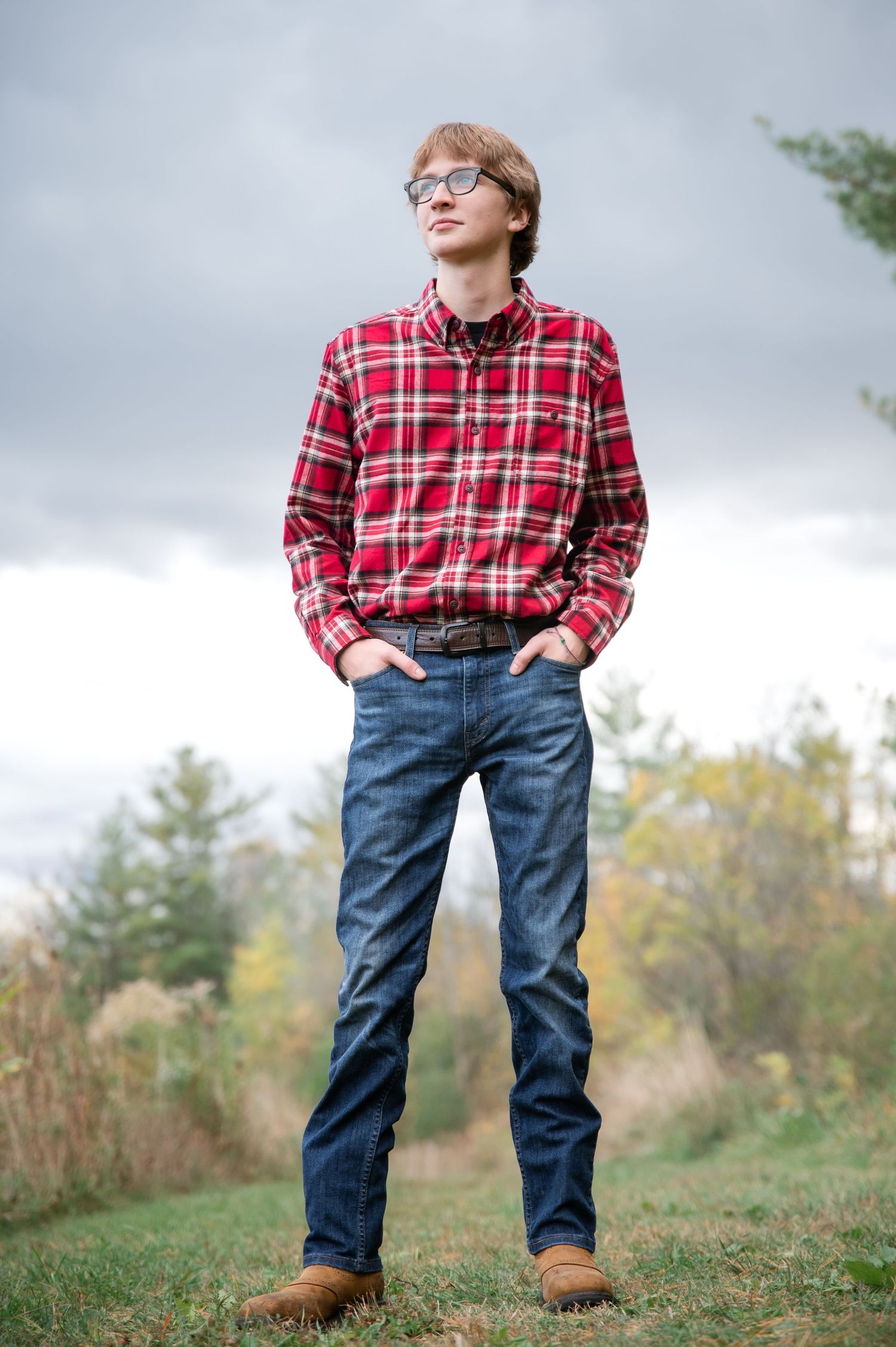 Person in red plaid flannel shirt and jeans stands on grassy hill against cloudy autumn sky.