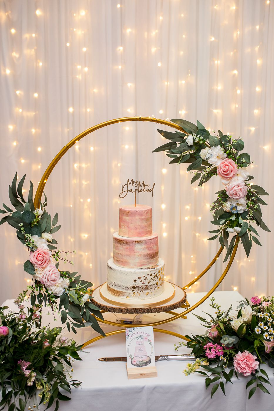 Elegant wedding cake display with fairy lights, pink roses and gold curved stands.