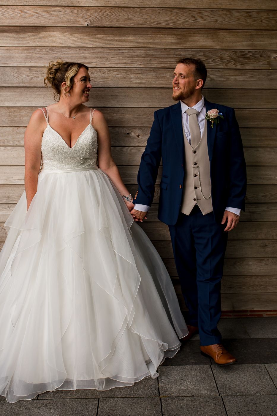 Wedding couple sits together against wooden wall backdrop, bride's white dress contrasting with groom's navy suit.