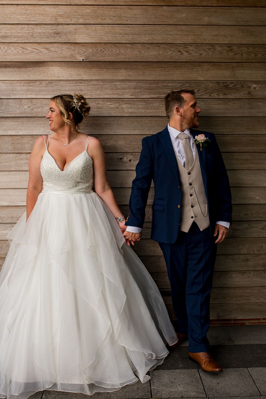Bride in flowing white gown and groom in navy suit pose against rustic wooden wall in artistic wedding photo series.
