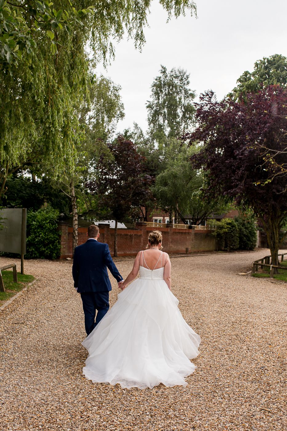 Couple walks hand in hand down tree-lined gravel path in wedding attire.