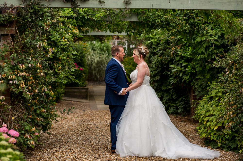 Wedding couple poses in lush garden setting with greenery and gravel path backdrop.