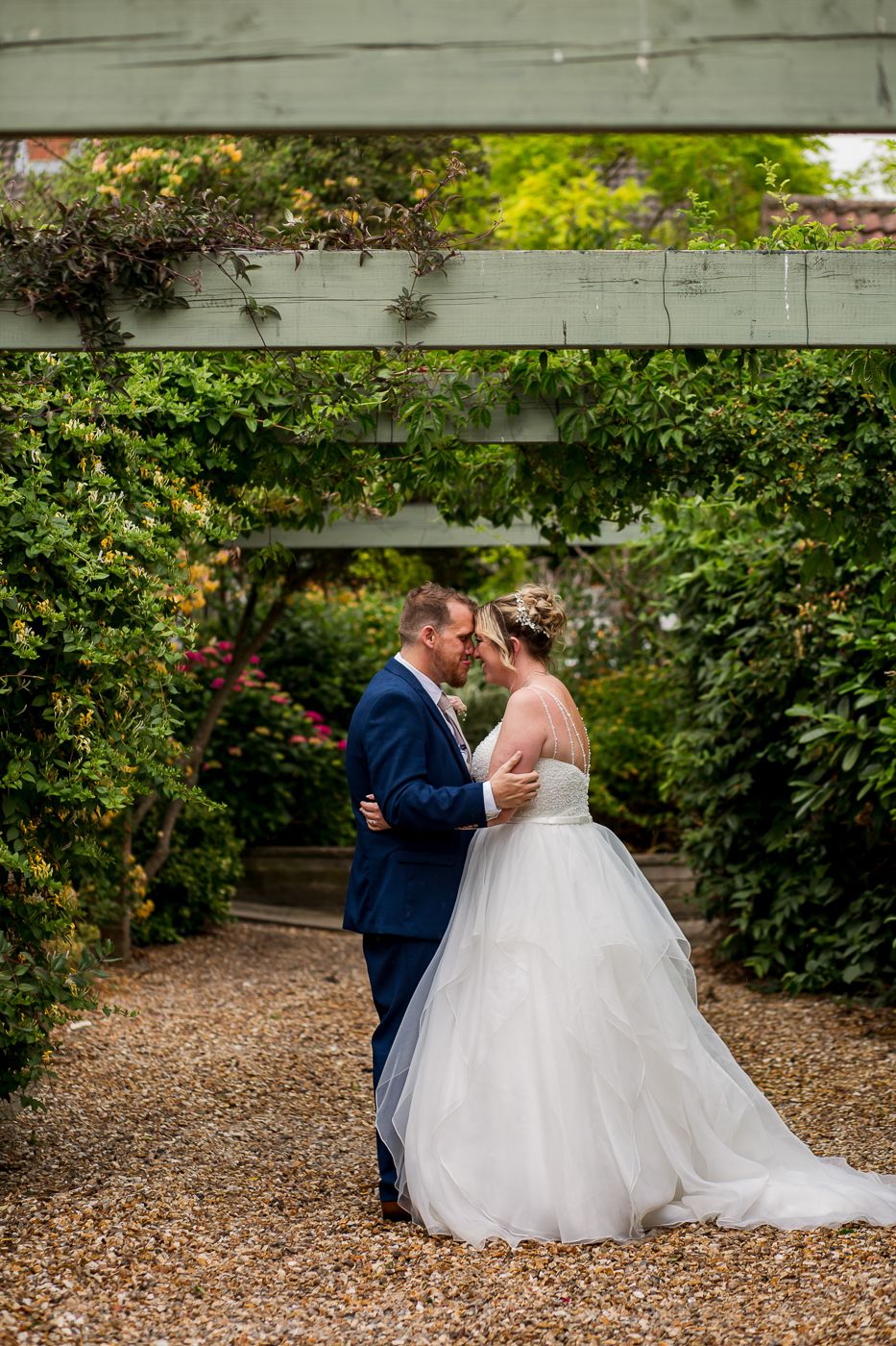 Wedding couple shares intimate moments in garden setting with leafy green backdrop and gravel path.