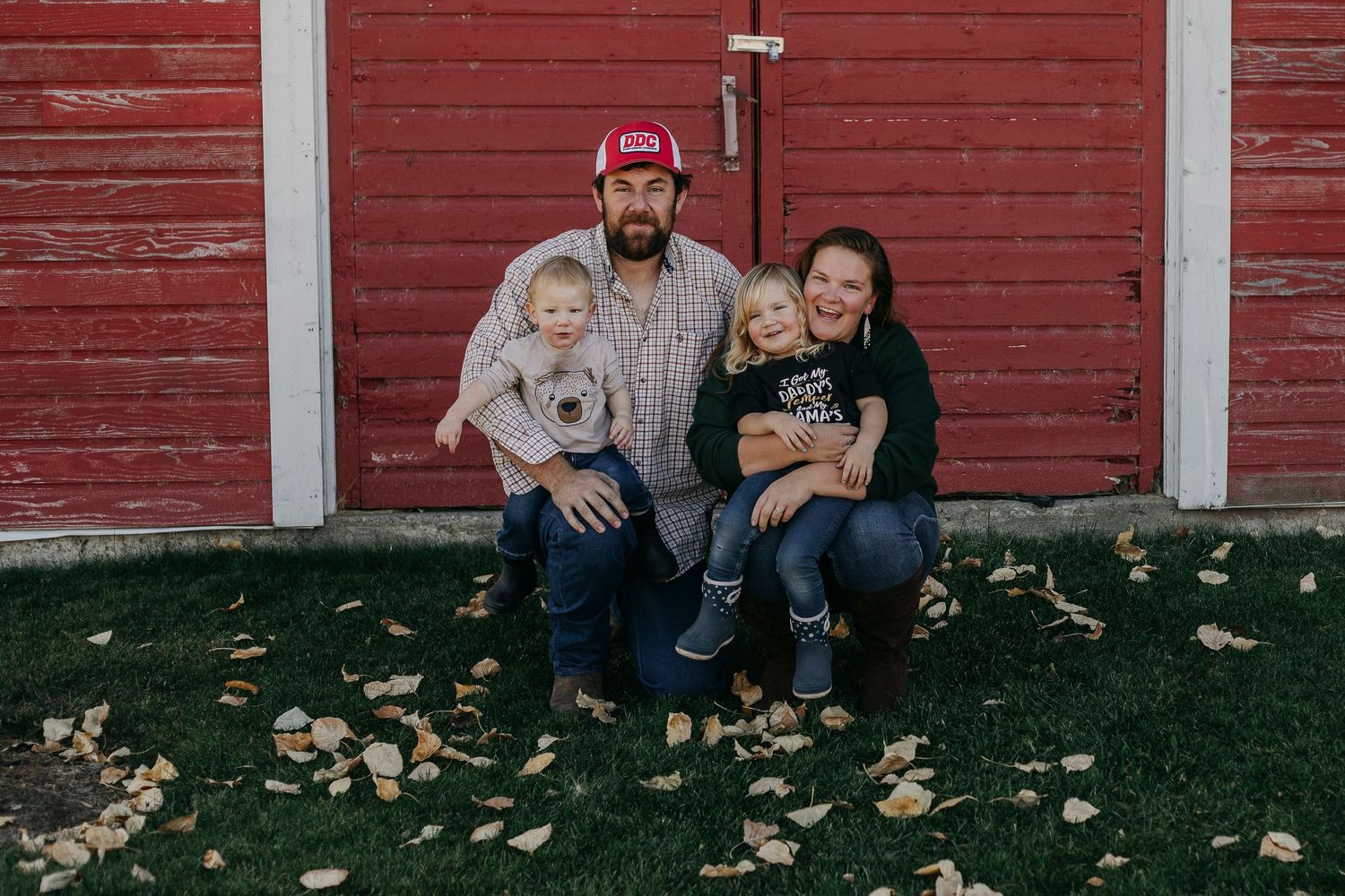 A family sits together on grass against a red barn wall during fall with autumn leaves scattered around them.