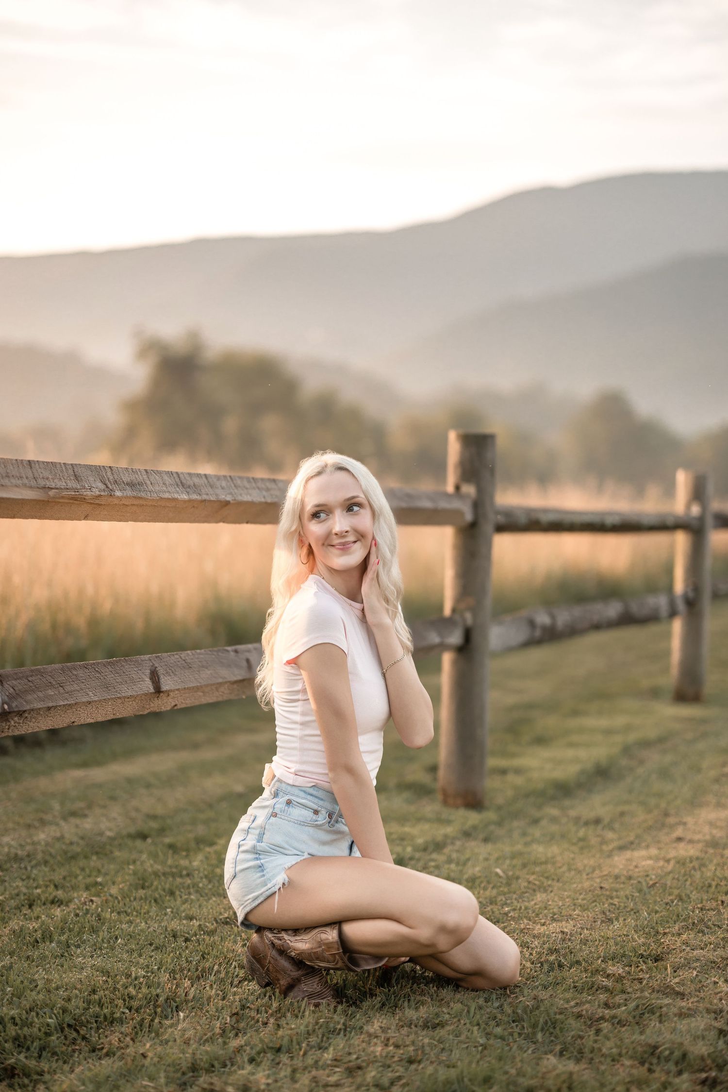 A woman in a white t-shirt and denim shorts poses on grassy ground near a wooden fence at sunset.