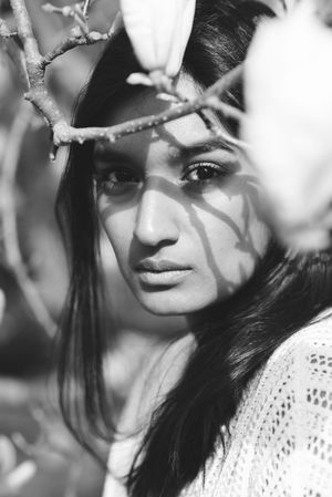 Artistic black and white portrait of a woman in a white knit sweater with shadows cast across her face from tree branches.