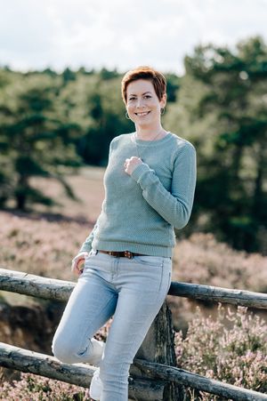 Person in light blue sweater sits casually on wooden fence surrounded by blooming heather in outdoor nature setting.