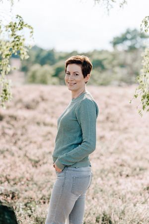 Person in a gray sweater and jeans poses casually in a sunlit outdoor field with soft natural lighting.