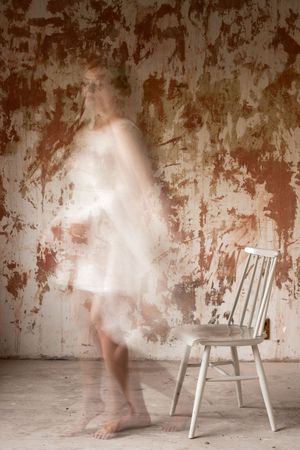Long exposure photograph shows ethereal blurred motion around a white wooden chair against a weathered wall.