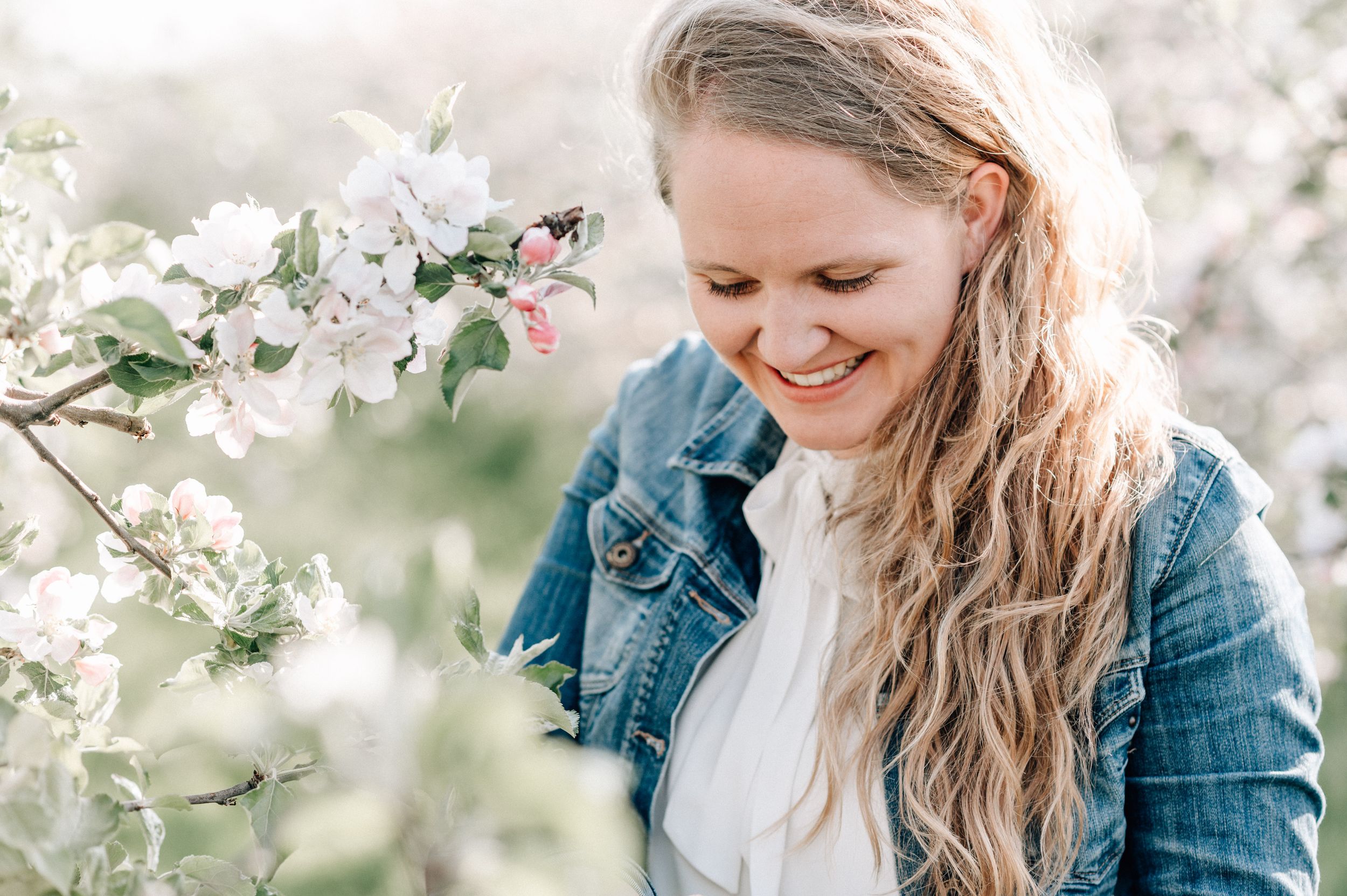 Someone in a denim jacket smiles while standing near blooming white spring blossoms in soft natural light.