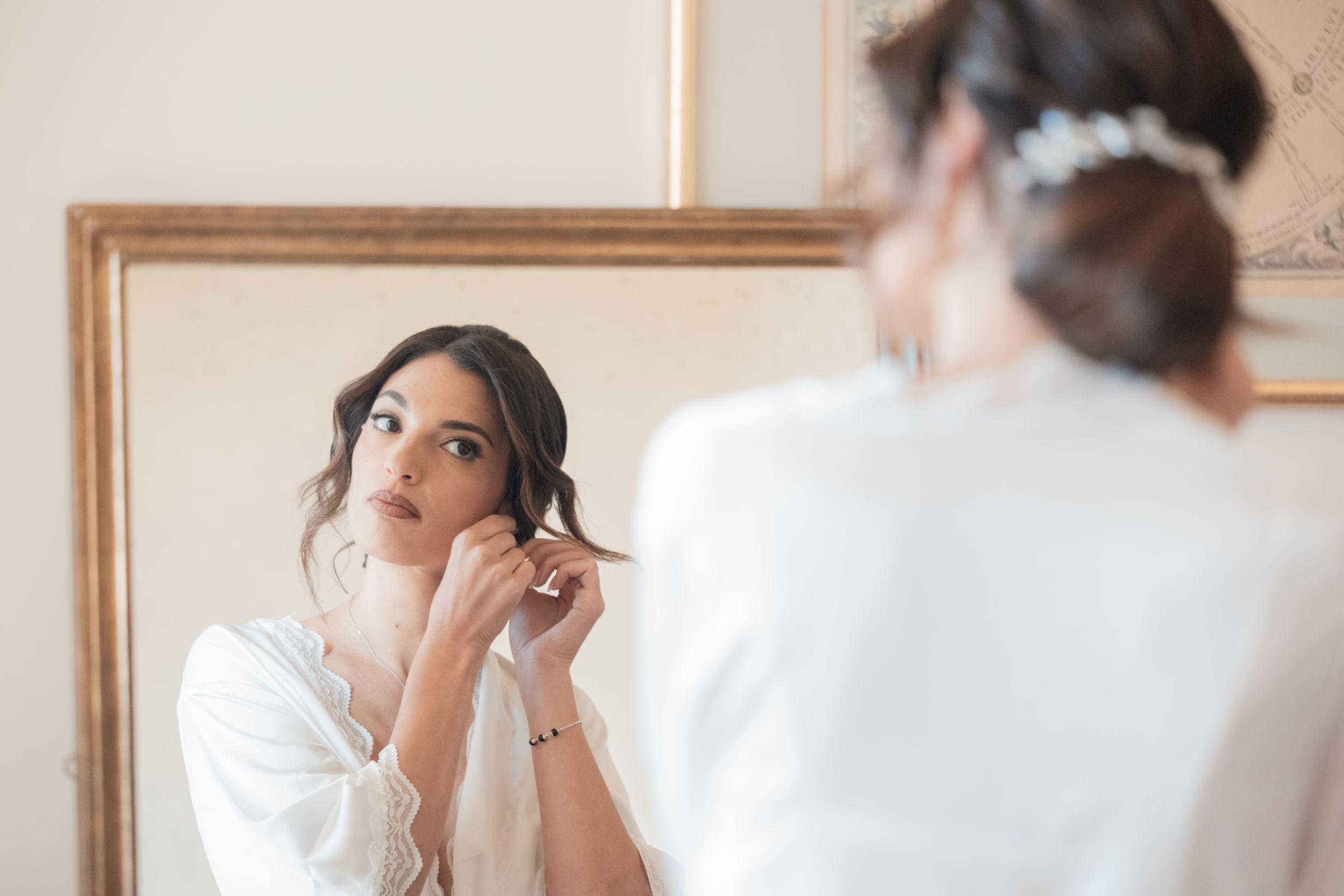 A bride in a white robe gets ready for her wedding day while sitting at a makeup vanity.
