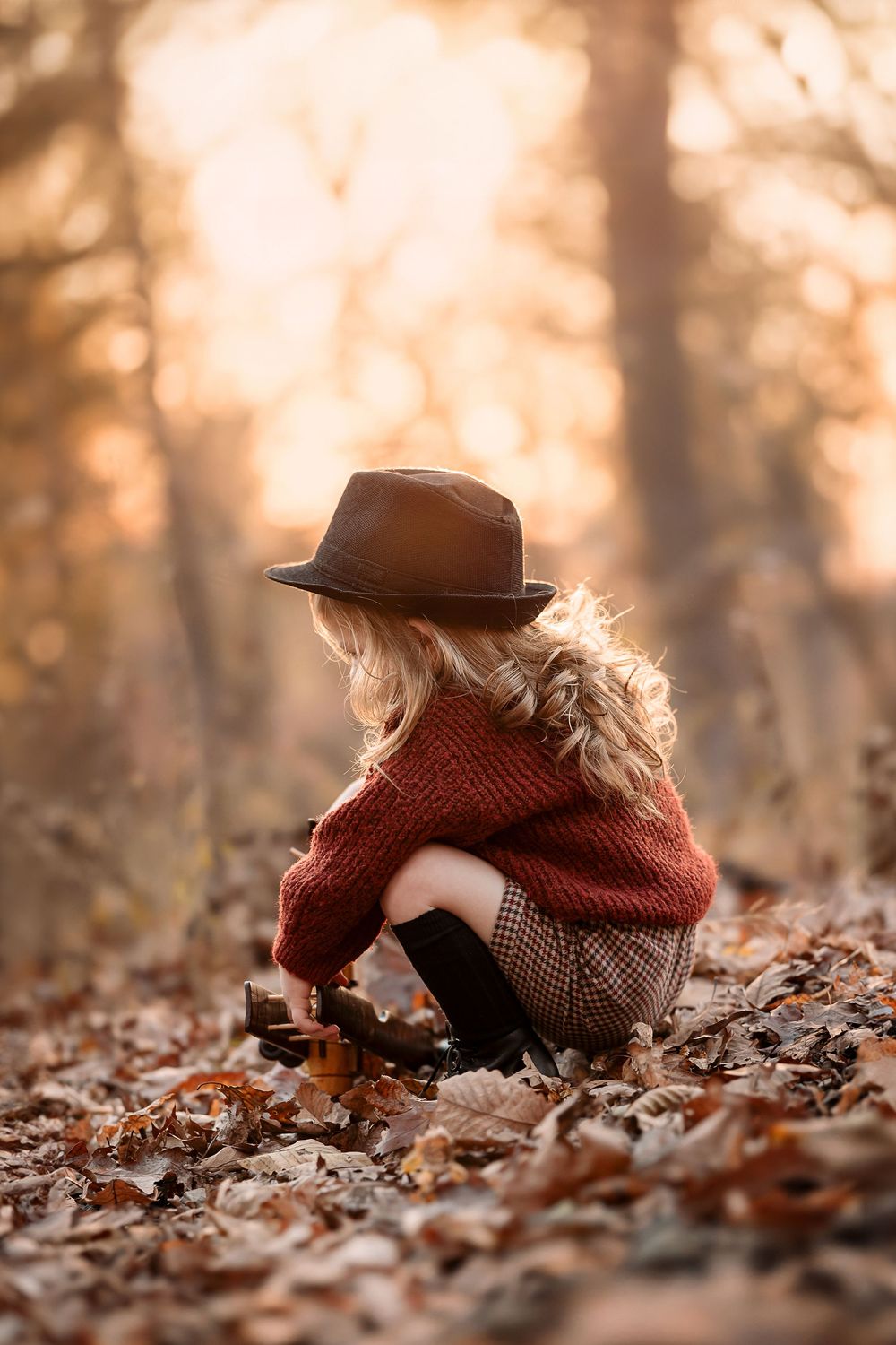Person in brown hat and scarf sits among fallen autumn leaves in warm golden sunlight.