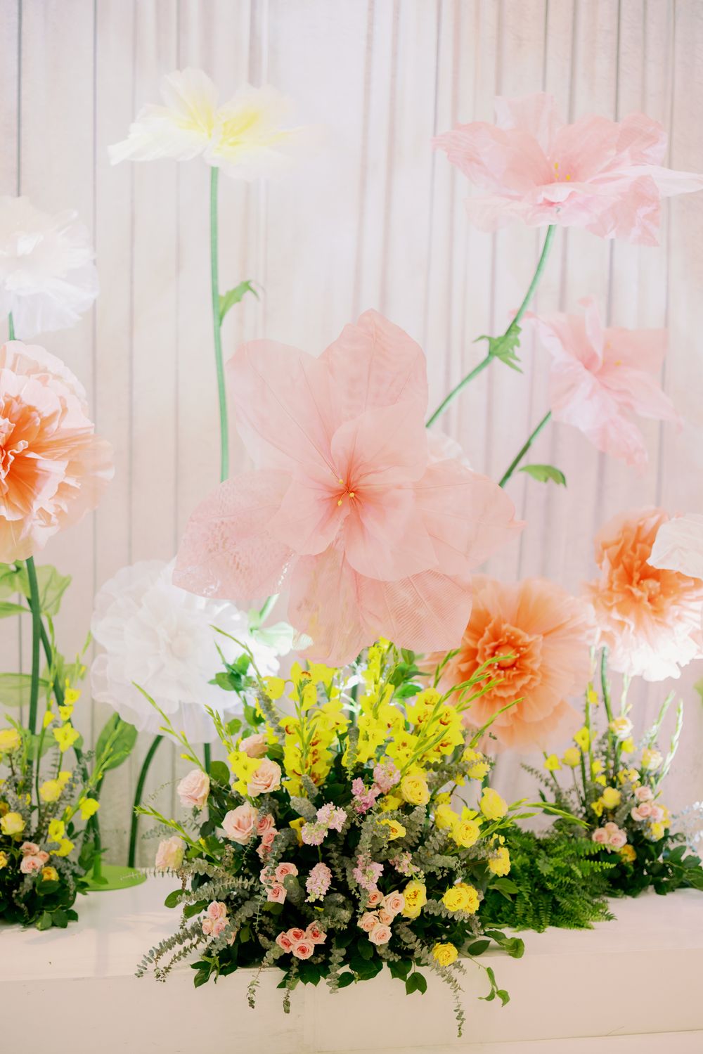 Delicate pink and peach paper flowers create an elegant backdrop with yellow and green floral arrangements below.