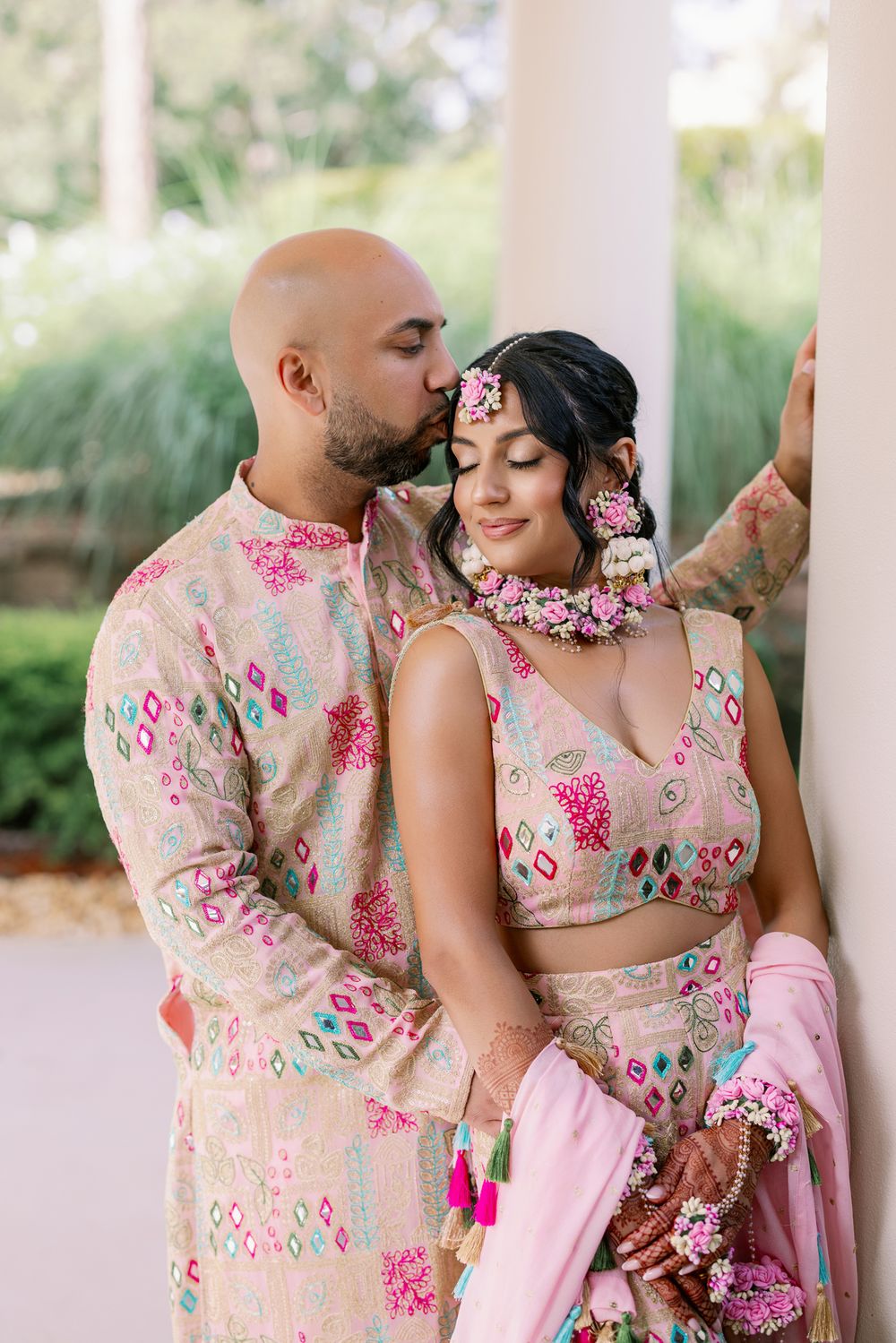 Couple wearing coordinated pink floral attire embrace tenderly against white pillars in a garden setting.